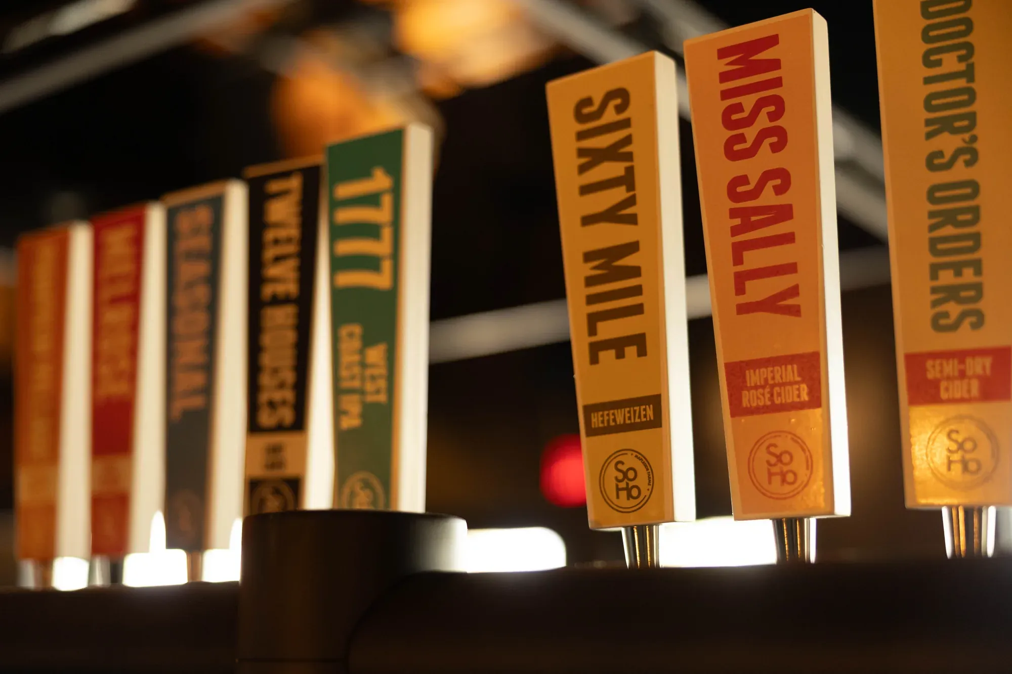 A row of colourful beer and cider tap handles, each with the SoHo monogram at the base
